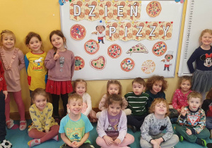 Dzieci ustawione na dywanie i pozują na tle dekoracji z okazji Światowego dnia pizzy.
