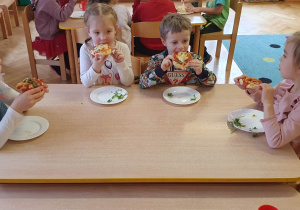 Dzieci siedzą przy stolikach i jedzą pizzę, którą wcześniej robiły.