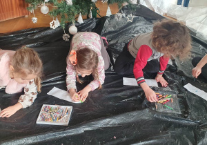 Dzieci siedzą na podłodze i malują kredkami oraz flamastrami wykonane przez siebie zakładki i okładki do książek.