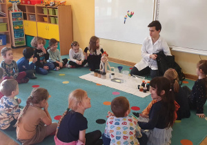 Dzieci siedzą na dywanie i słuchają prowadzącego warsztaty chemiczne LABOLO, który zapoznaje je z tematem zajęć.