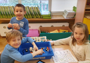 Chłopcy siedzą z dziewczynką przy stole i układają żuka z klocków lego na zajęciach z robotyki.