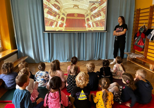 Dzieci siedzą na podłodze i słuchają prowadzącej warsztaty nt. opery.