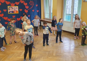 Dzieci wspólnie z trenerką zumby rozciągają mięśnie szyi kręcąc głową i trzymając ręce na biodrach.