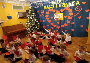 Dzieci elegancko ubrane siedzą na podłodze i trzymają w rękach serduszka , którymi ruszają w rytmie piosenki na występie z okazji Dnia Babci I Dziadka.