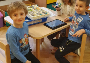 Dwóch chłopców z dziewczynką siedzą przy stole i prezentują swoją pracę wykonaną z klocków lego na zajęciach z robotyki.