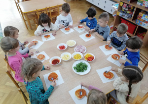 Dzieci siedzą przy stolikach i smarują spód pizzy sosem pomidorowym.