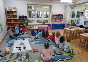 Dzieci siedzą na dywanie i obserwują jak prowadzący warsztaty chemiczne LABOLO wykonuje doświadczenie.
