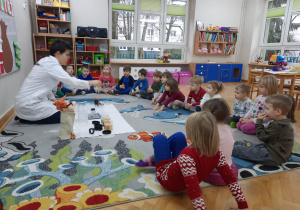 Dzieci siedzą na dywanie i obserwują jak prowadzący warsztaty chemiczne LABOLO pokazuje im co znajduje się w pojemniczku.