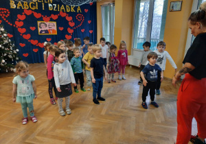 Dzieci stoją przed trenerką i powtarzają za nią ruchy, stojąc w lekkim rozkroku unosząc ramiona do góry.
