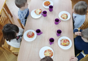 Dzieci siedzą przy stolikach i przyglądają się wykonanym przez siebie pizzą z okazji Światowego Dnia Pizzy.