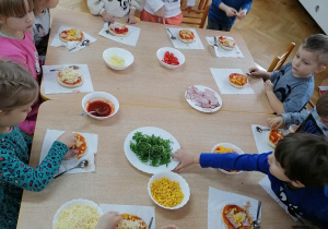 Dzieci siedzą przy stolikach i nakładają różne składniki na swoją pizzę.