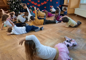 Dzieci leżą na podłodze i wspólnie z trenerką zumby wykonują ćwiczenia rozciągające.