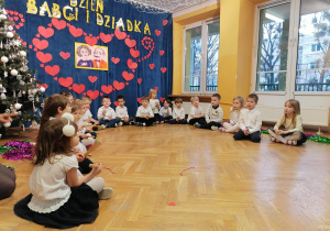 Dzieci siedzą na podłodze w półkolu śpiewają piosenkę i grają na instrumentach.