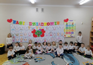 Dzieci siedzą na dywanie elegancko ubrane, a w tle wiszą portrety babć i dziadków.