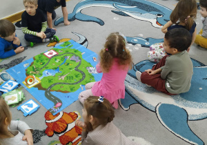 Dzieci siedzą w kole na dywanie, wspólnie próbują wymyśleć trasę robocika ślimaka, którą będzie się poruszał po macie.