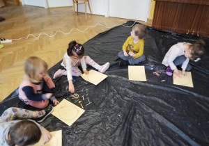 Dzieci siedzą na podłodze i samodzielnie próbują narysować poszczególne części buzi na szablonie głowy.