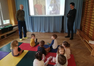 Dzieci siedzą na materacach i oglądają prezentację na temat portretów na warsztatach Fundacji Szkatułka.