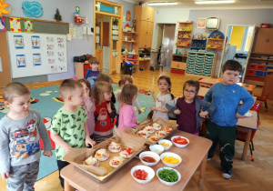 Dzieci stoją dookoła stolika i prezentują swoje pizze, które przygotowali z okazji Światowego Dnia Pizzy.