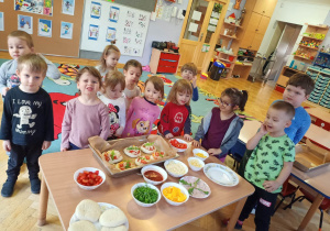 Dzieci stoją dookoła stolika i prezentują swoje pizze, które przygotowali z okazji Światowego Dnia Pizzy.
