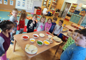 Dzieci stoją dookoła stolika i zapoznają się ze składnikami, które będą mogli dołożyć do swojej pizzy.