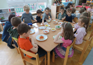 Dzieci siedzą przy stolikach i dodają różne składniki do swojej pizzy.