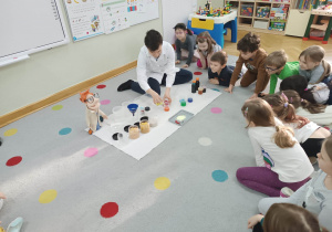 Dzieci siedzą na dywanie w kole i obserwują jak prowadzący warsztaty chemiczne LABOLO miesza ze sobą 3 kolorowe ciecze.