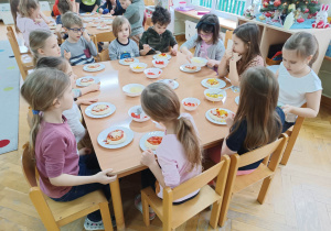 Dzieci siedzą przy stolikach i dodają różne składniki do swojej pizzy.