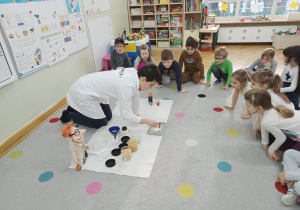 Dzieci siedzą na dywanie w kole i obserwują jak prowadzący warsztaty chemiczne LABOLO wykonuje doświadczenie.