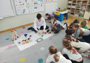 Dzieci siedzą na dywanie w kole i obserwują jak prowadzący warsztaty chemiczne LABOLO miesza ze sobą 3 kolorowe ciecze.
