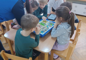 Dzieci siedzą przy stoliku i z pomocą prowadzącej warsztaty robotyki próbują złożyć robota z klocków lego.
