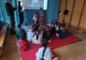 Dzieci siedzą na materacach i obserwują jak dwoje dzieci przebranych w kostiumy aktorów z opery odgrywają scenkę.