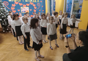 Dzieci stoją ustawione w kilku rzędach elegancko ubrane podczas występu dla dziadków.