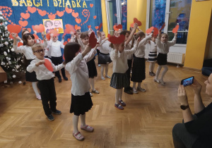 Dzieci ustawione w kilku rzędach elegancko ubrane śpiewają piosenkę dla dziadków wymachując papierowymi serduszkami nad głowami.