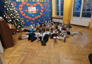 Dzieci leżą w kole na brzuchach, machając nogami podczas występu z okazji dnia babci i dziadka.