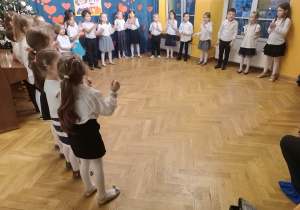 Dzieci stoją ustawione w półkolu recytują wiersz dla dziadków z okazji ich święta klaszcząc w dłonie.