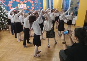 Dzieci elegancko ubrane tańczą w parach trzymając się za ręce tworząc koło podczas występu dla dziadków.