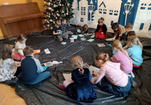 Dzieci siedzą na podłodze i malują kredkami i flamastrami wykonane przez siebie okładki do książek.