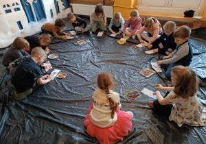 Dzieci siedzą w kole na podłodze i malują według własnego pomysłu zakładki do książek.