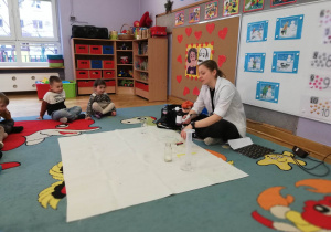 Dzieci siedzą na dywanie i obserwują prowadzącą wykonującą doświadczenie związane z ciśnieniem.