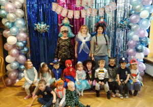 Dzieci przebrane w różne stroje wspólnie z wychowawczynią pozują do zdjęcia grupowego na tle dekoracji z balu karnawałowego.