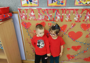 Chłopiec z dziewczynką ubrani na czerwono trzymają się za rączki i pozują do zdjęcia na tle napisu Walentynki.