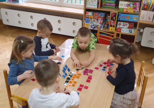 Dzieci siedzą przy stoliku, na przezroczystej folii przyklejają kolorowe kawałki folii tworząc z nich witraż.