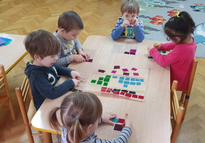 Dzieci siedzą przy stoliku, na przezroczystej folii przyklejają kolorowe kawałki folii tworząc z nich witraż.