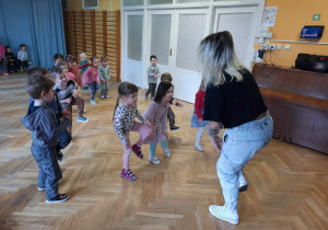 Dzieci stoją w rozsypce na sali gimnastycznej i powtarzają ruchy taneczne za trenerką zumby podnosząc jedną nogę do góry.