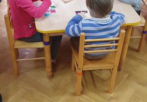 Dzieci siedzą przy stoliku, na przezroczystej folii przyklejają kolorowe kawałki folii tworząc z nich witraż.
