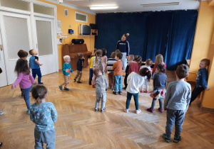 Dzieci stoją w rozsypce na sali gimnastycznej i powtarzają ruchy taneczne za trenerką zumby machając głową na boki.