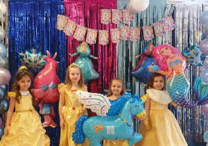Dziewczynki przebrane za Bellę pozują do zdjęcia z dużymi balonami postaci z bajek na tle dekoracji z balu karnawałowego.
