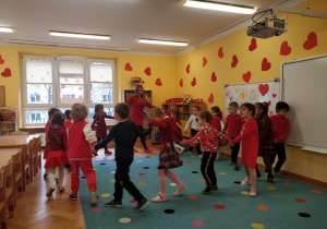 Dzieci ubrane na czerwono ustawione w kole tańczą z wychowawczynią do ulubionej muzyki.