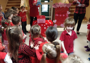 Dzieci ubrane na czerwono stoją i słuchają jak Pani dyrektor z nauczycielką wyczytują imiona dzieci, które otrzymają walentynkę.