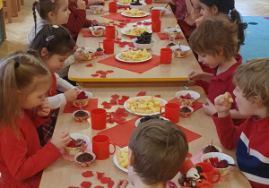 Dzieci ubrane na czerwono siedzą przy stolikach udekorowanych na czerwono i jedzą walentynkowy poczęstunek.